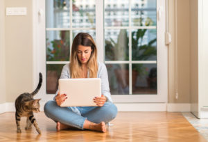 A young woman sits on a wood floor next to a striped cat, smiling at laptop screen