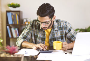 Man holding yellow mug uses calculator while facing a laptop